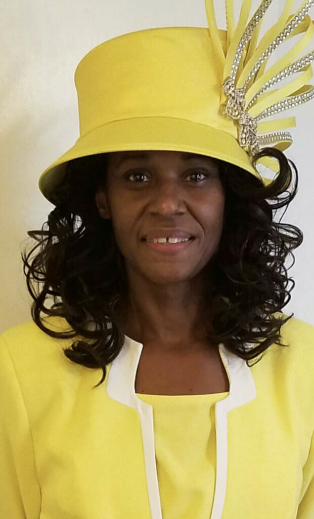 A smiling black woman with long, wavy hair, wearing a yellow suit jacket, white necklace and a matching yellow hat.
