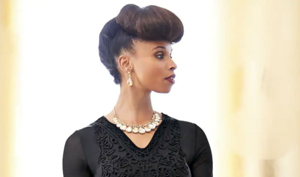 An African-American woman with a fancy updo hairstyle, wearing a black V-neck top and a pearl necklace.