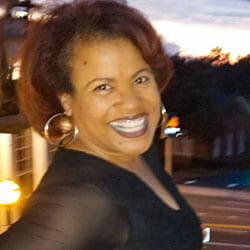 Leslie, a smiling African-American woman with short copper hair, wearing a black V-neck top and large hoop earrings.