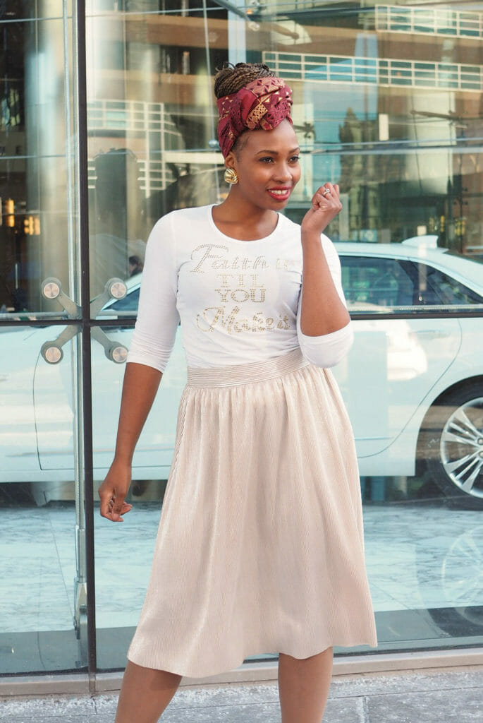 A black woman in a 'Faith it til You Make it' white T-shirt and a light pink pleated skirt, on a city sidewalk.