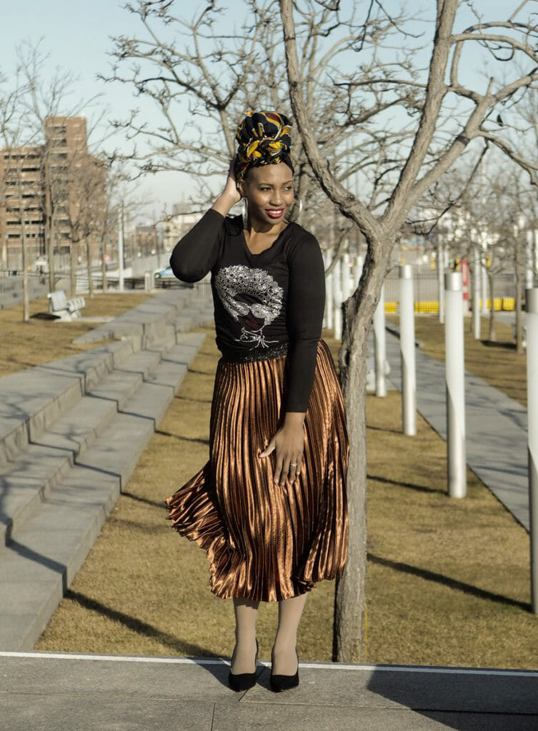 A black woman in a city, wearing a black, long sleeve t-shirt with a lady head applique, and a bronze pleated skirt.