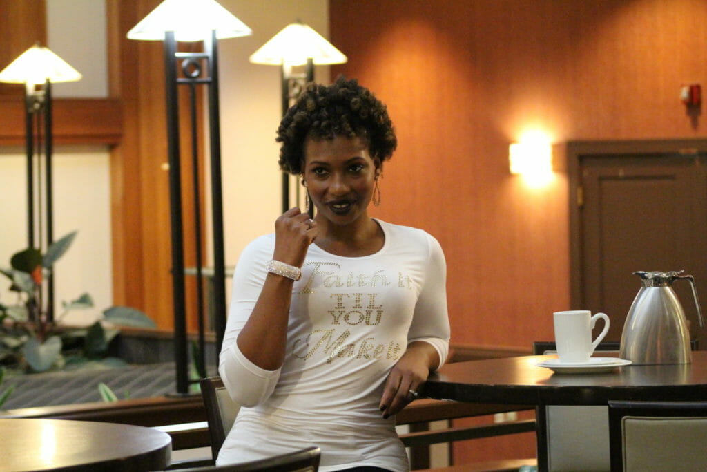 An African-American woman at a cafe, wearing a 'Faith it til You Make it' white T-shirt.