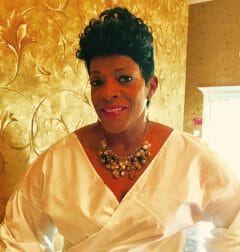 An African-American woman in a silky, pale yellow wrap blouse and bib necklace, in front of a gold wall.