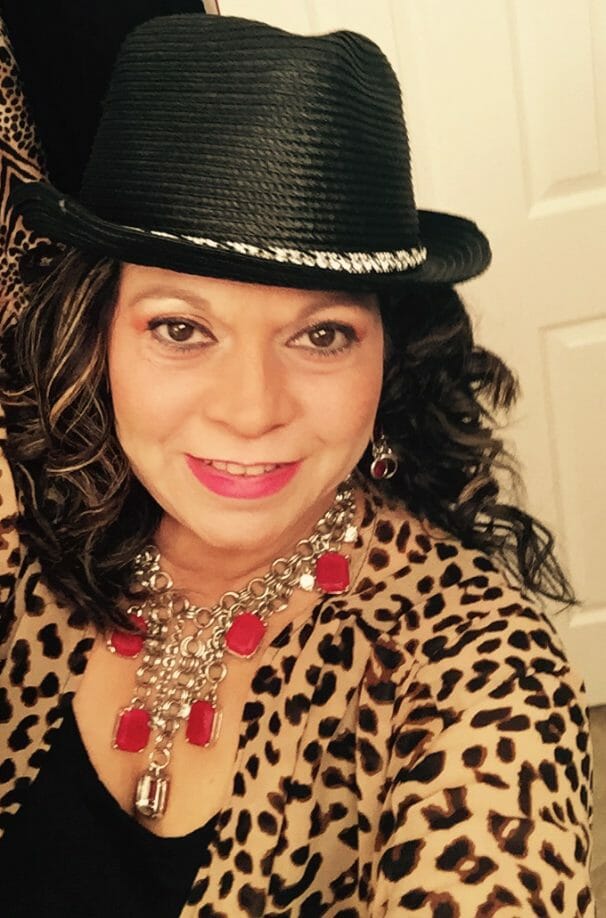 A smiling African-American woman wearing a leopard print top, red necklace and a black hat.