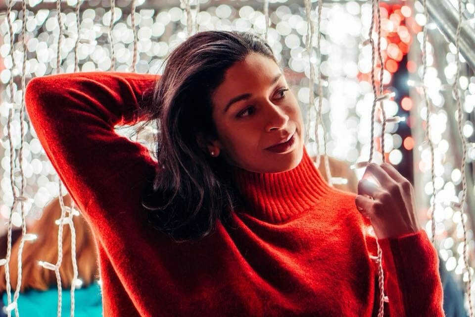 A black woman with shoulder-length hair wearing a red sweater, in front of white string lights.