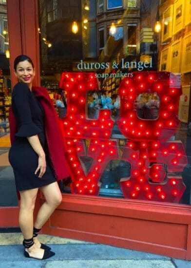 A black woman in a black dress standing in front of a storefront window with a red, lit LOVE sign.
