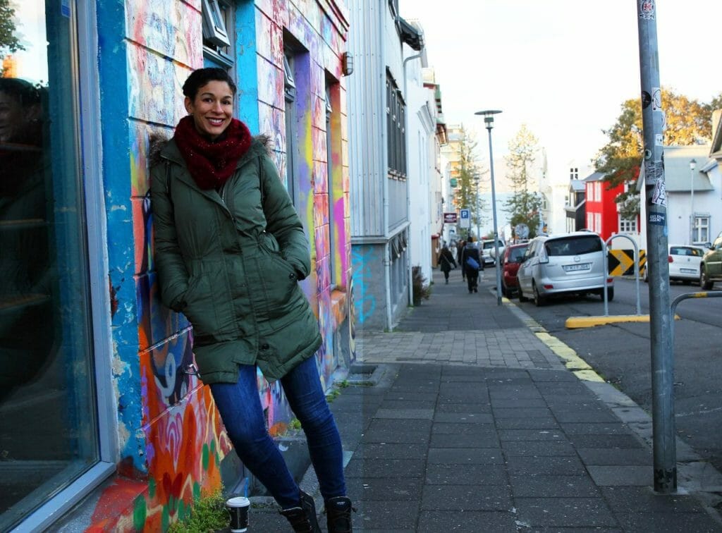 An African-American woman in a winter coat and jeans, leaning against a graffiti wall on a city sidewalk.