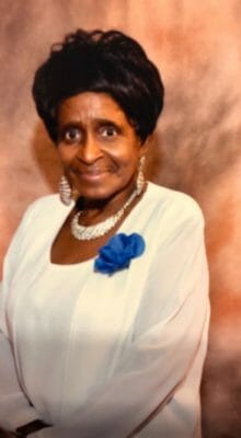 Customer Agnes P., a smiling black woman wearing a white jacket dress, with white pearls and a blue flower corsage.