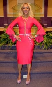 Customer Synetra, a smiling black woman with blonde hair, wearing a long-sleeved bright pink dress.