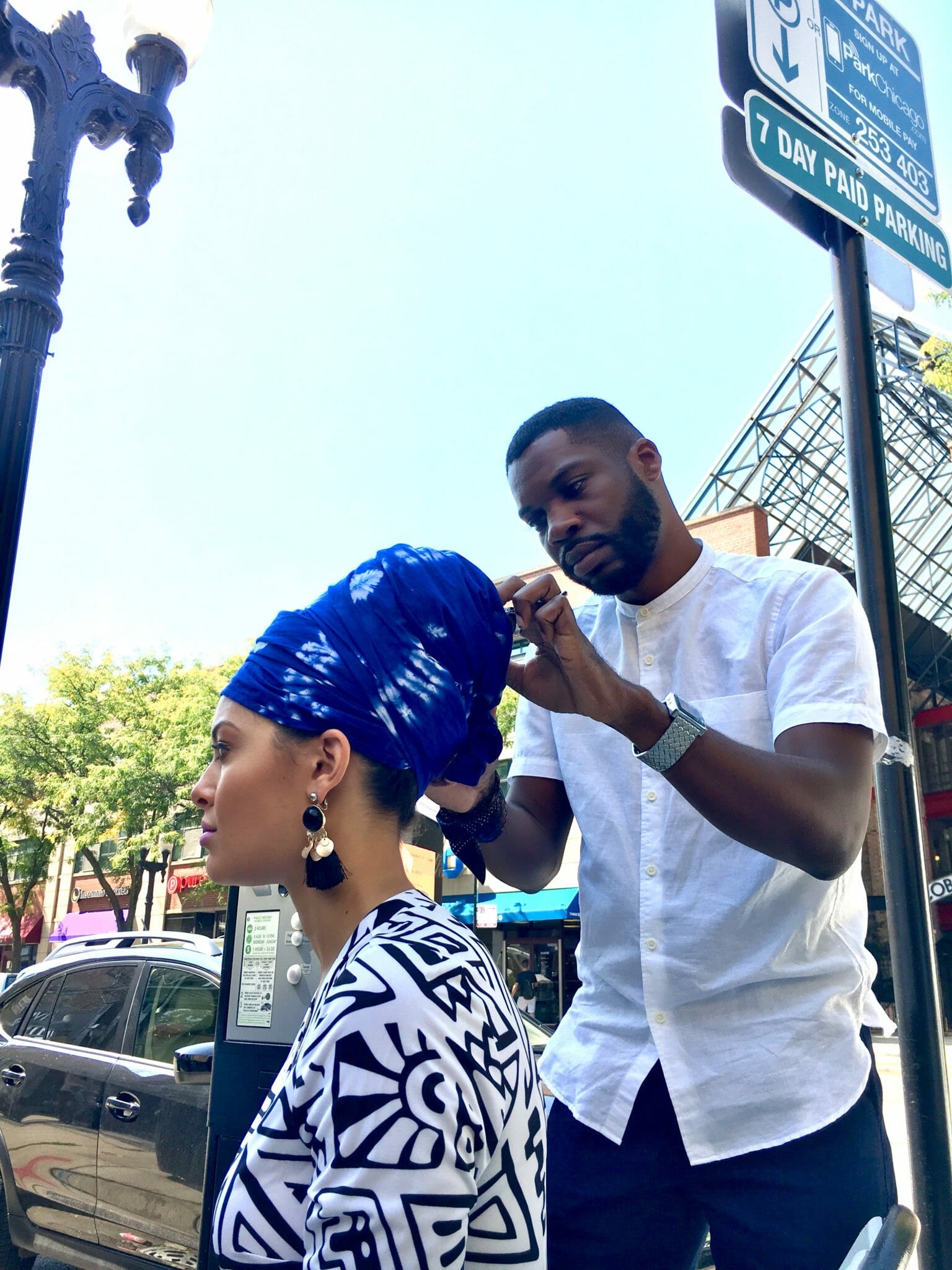A Black man in a white shirt adjusting a blue headwrap on a black woman.