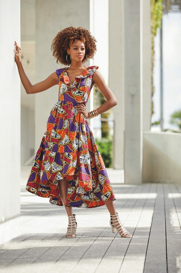 A black woman in a purple, white and orange multi-print Afrocentric dress with a flared skirt, and a matching headwrap.