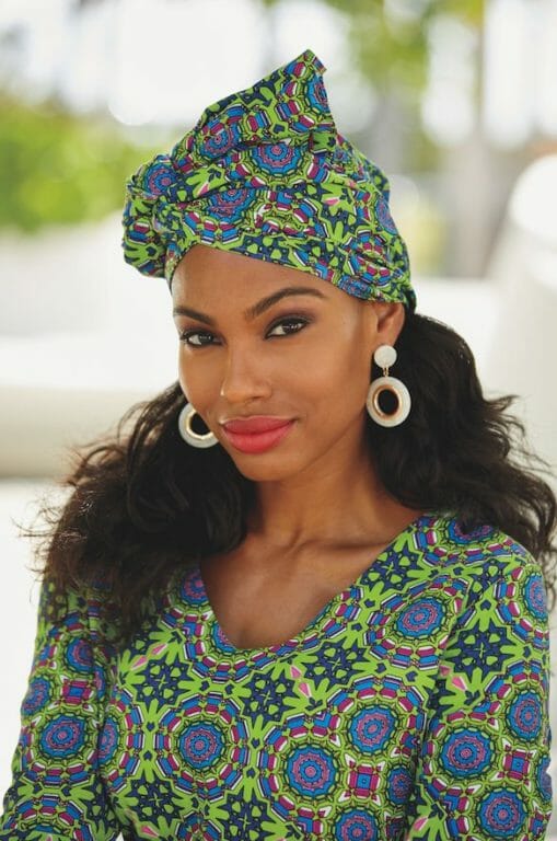 A black woman with long wavy hair and white hoop earrings wearing a green and blue Afrocentric headwrap.
