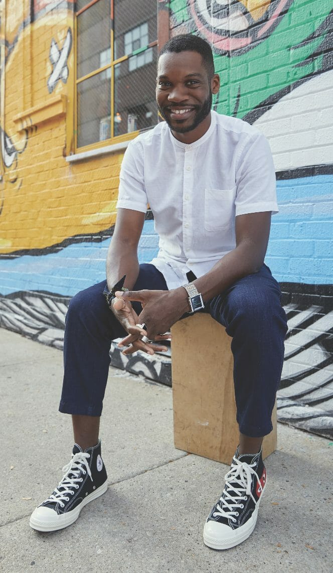 A black man in a white shirt and jeans sitting on a wood block in front of a graffiti wall.