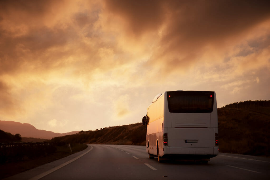 A white bus driving forward on a road, with a sunset view.