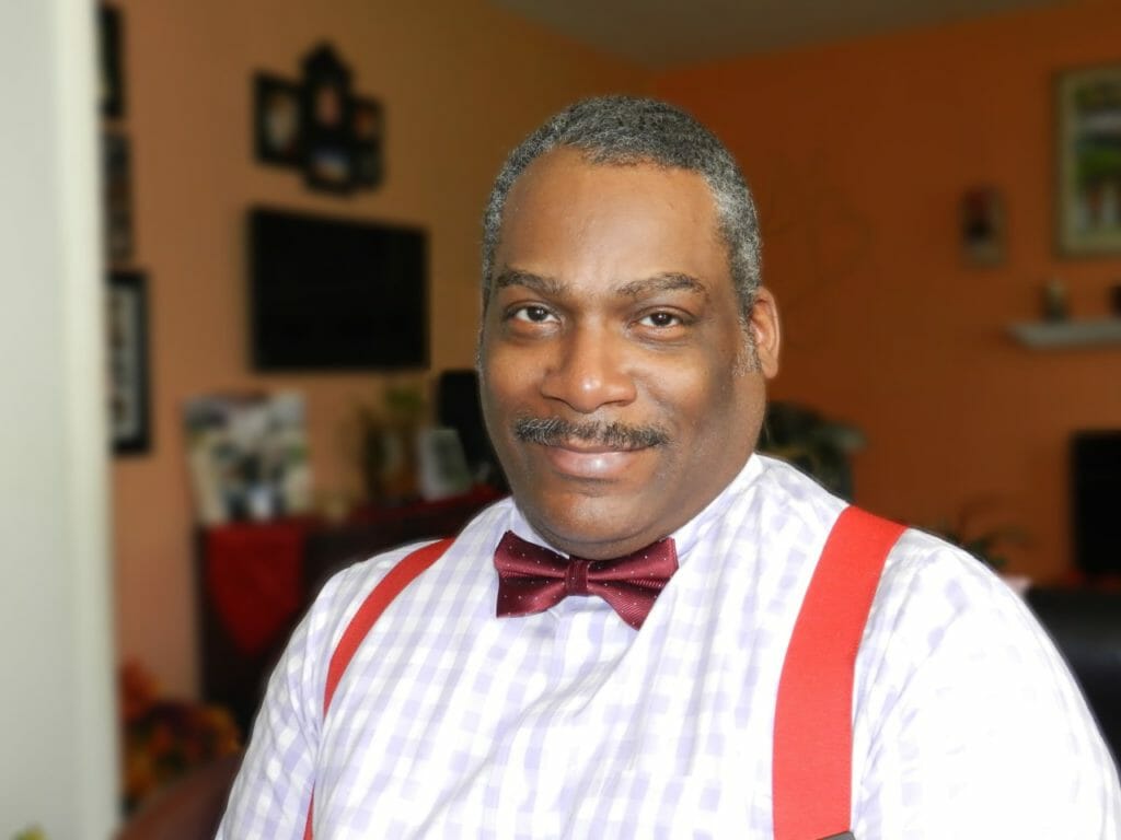 A black man wearing a white dress shirt with a red bow tie and red suspenders.