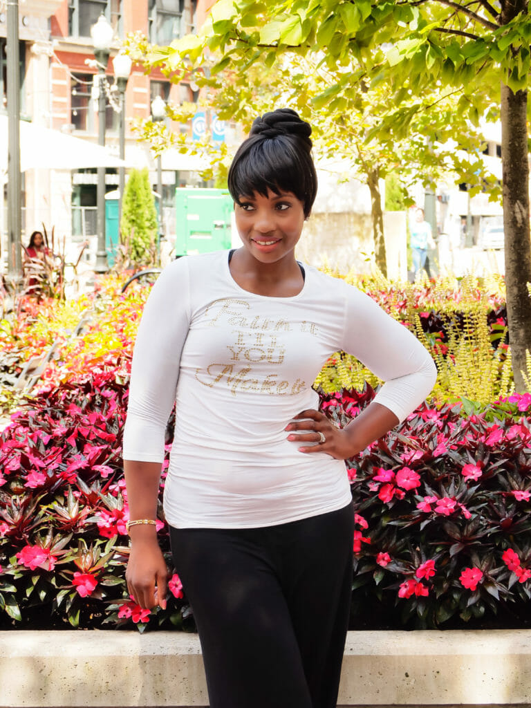A black customer wearing a white top, black slacks, and hair in a high bun with bangs, in front of blooming flowers.