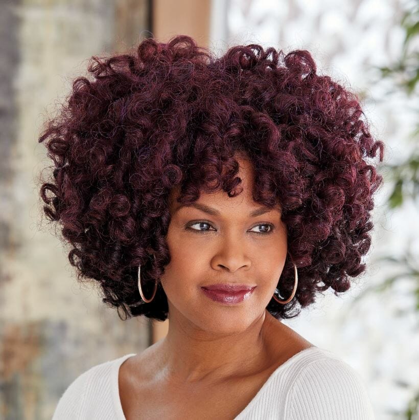 A smiling African-American woman wearing a white top and a full, curly burgundy Vivica Fox wig with bangs.