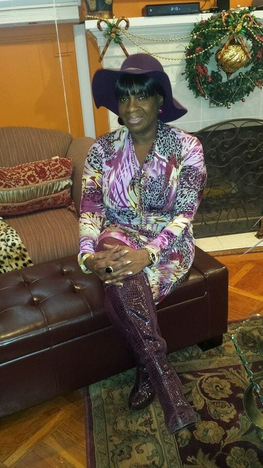 Ashro Woman of the Week, Beverly, a black woman, wearing a lavender print dress, brown boots, and brimmed hat.