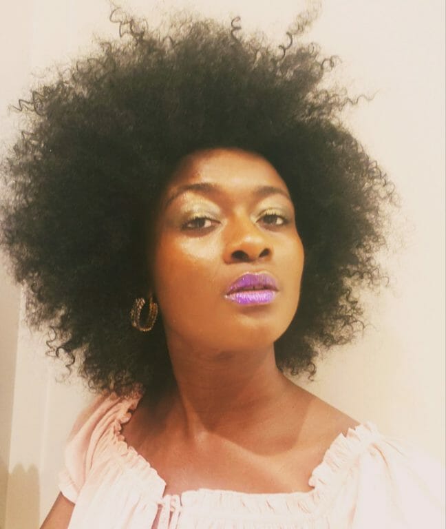 An African-American woman with a full Afro hairdo, wearing a soft pink top, earrings and pink lipstick.
