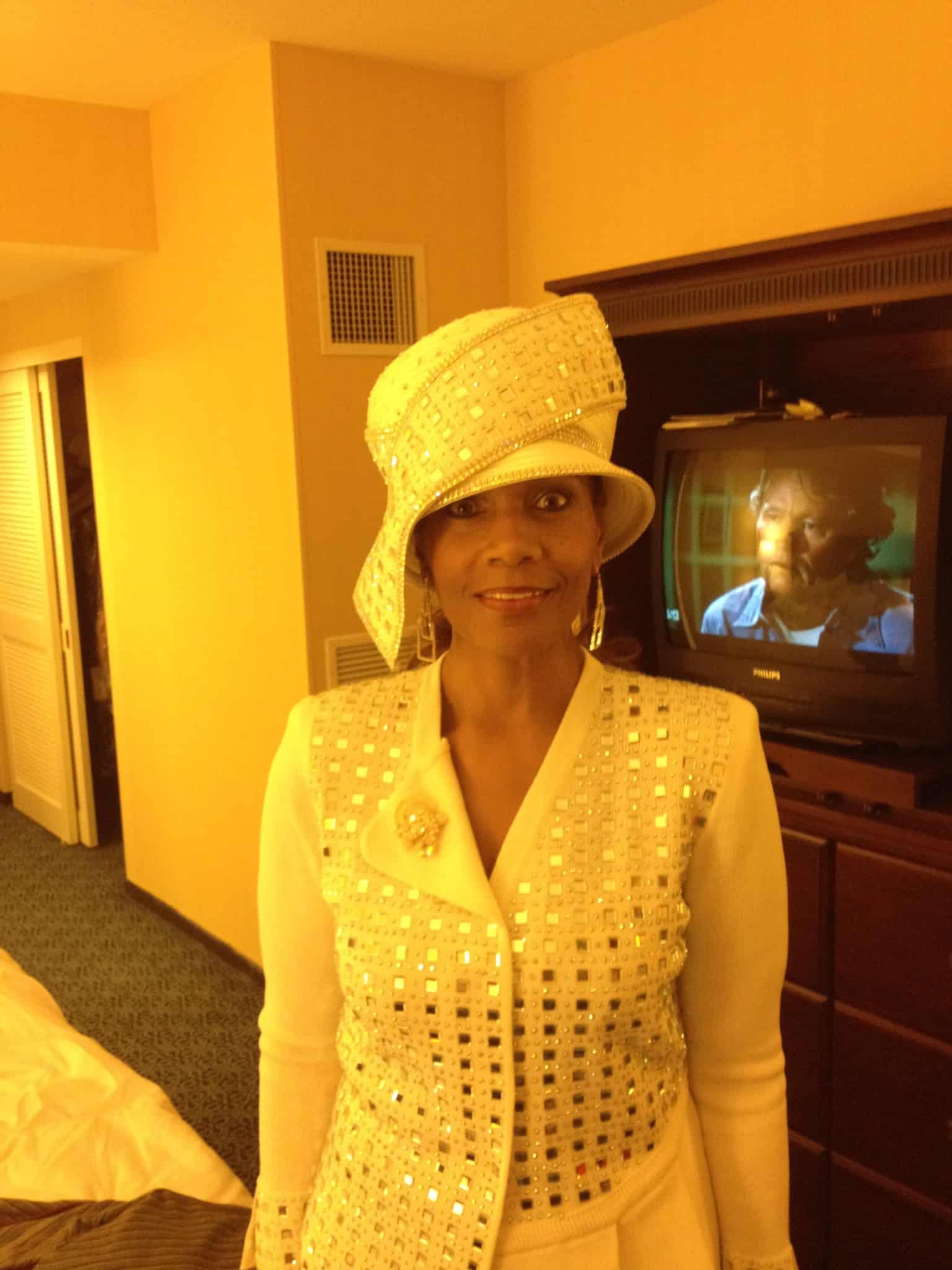 Ashro Woman of the Week, Dr. Angela, a black woman in a cream hat with gold polka dots and matching suit.