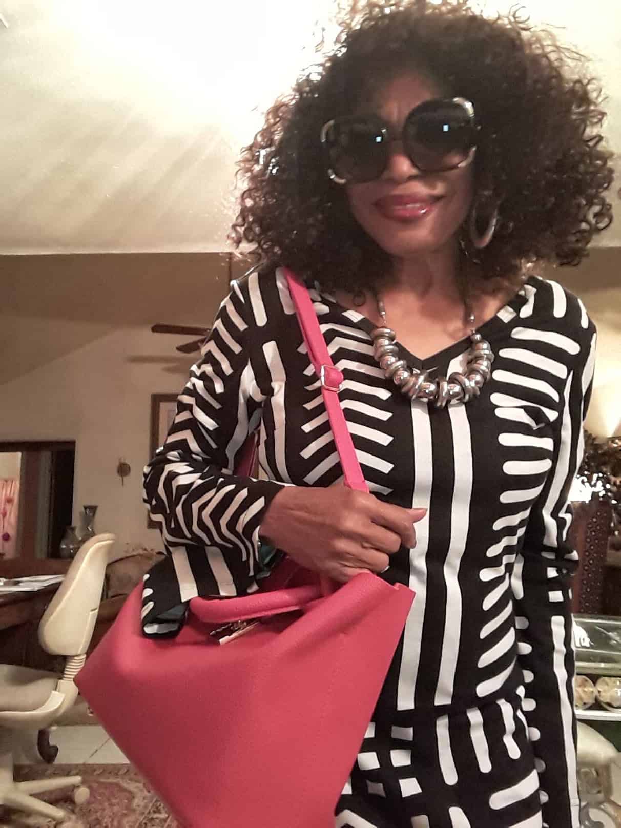 Ashro Woman of the Week, Lucy, a black woman in a black and white dress, sunglasses, with a coral shoulder bag.