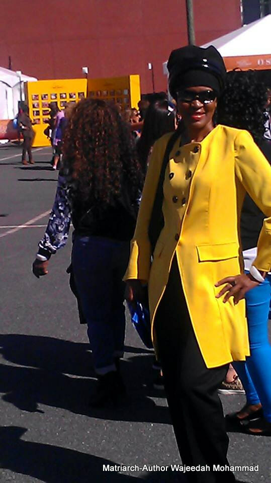 Ashro Woman of the Week WAJEEDAH, a black woman in sunglasses, long yellow jacket, black pant, and a black headwrap.