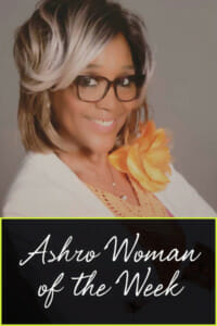 Ashro Woman of the Week CAROLYN, a smiling blonde black woman in glasses wearing a white jacket with flower.