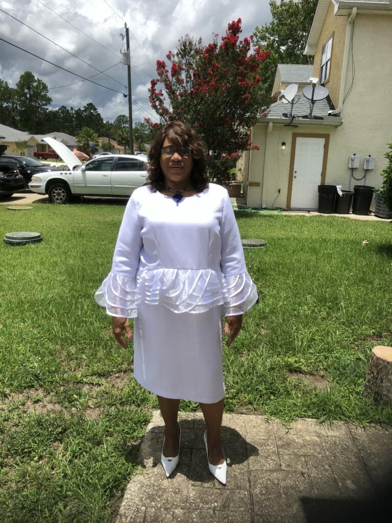 FRANCES LARRY, an African-American woman in a white dress with a peplum and flared sleeves.