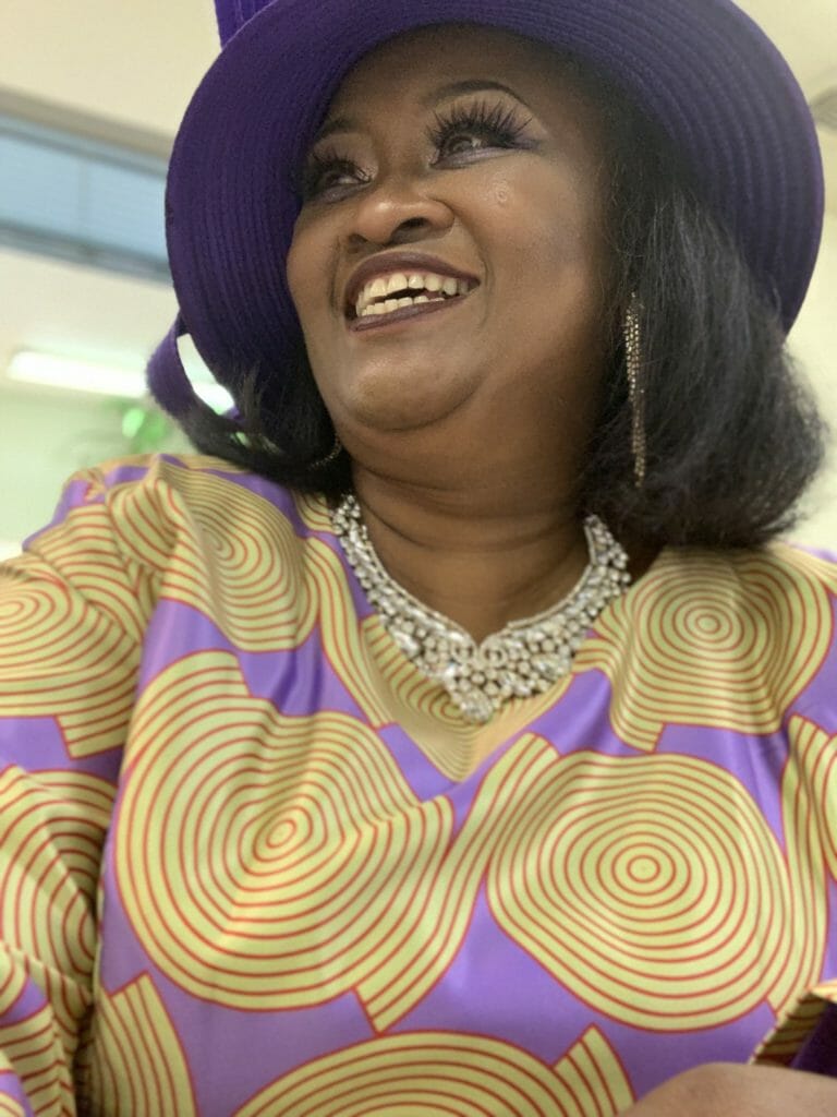 RENEE W., a smiling black woman in a yellow and lavender print dress with a bead necklace and black hat.