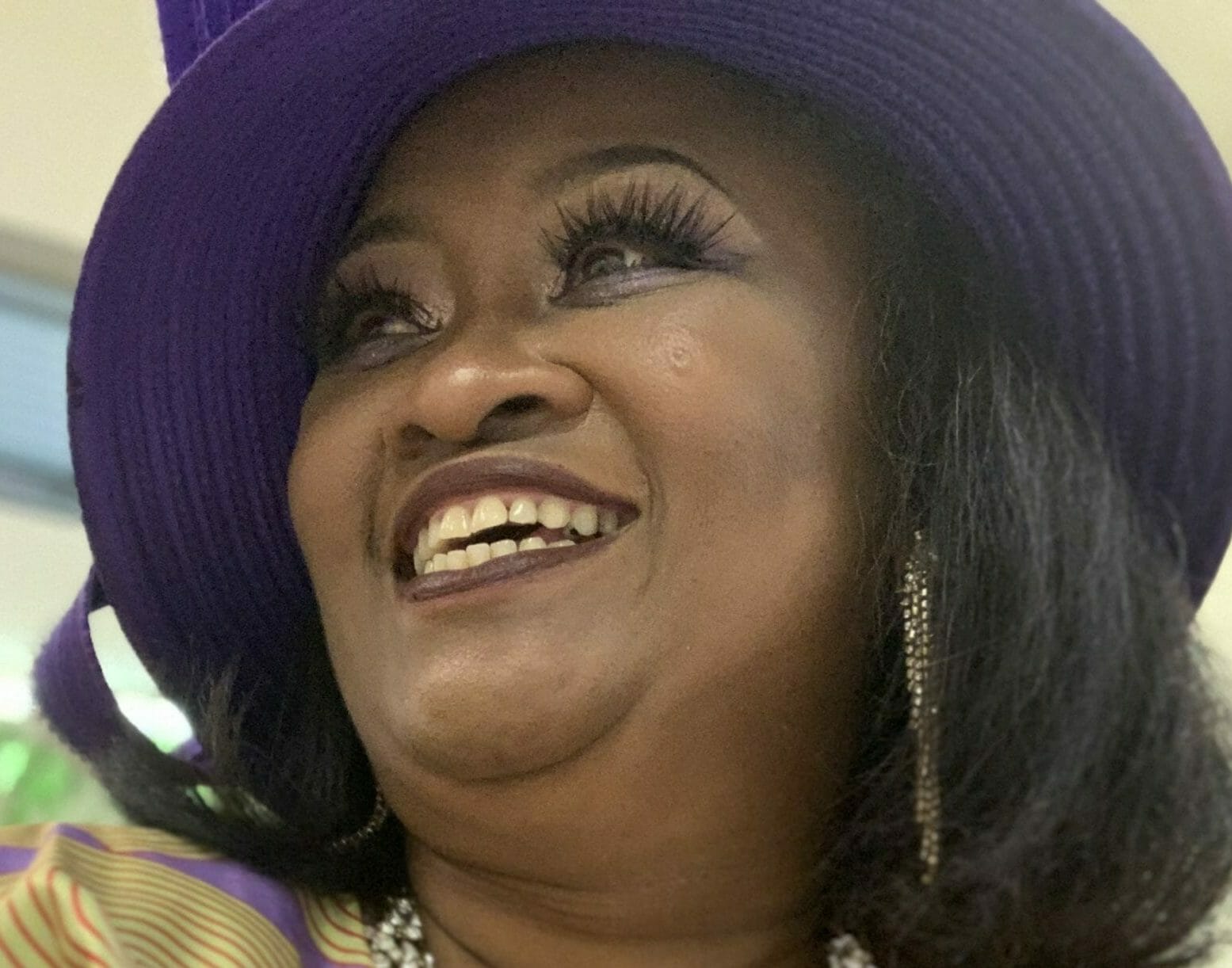 RENEE W., a smiling black woman in a yellow and lavender print dress with a bead necklace and black hat.