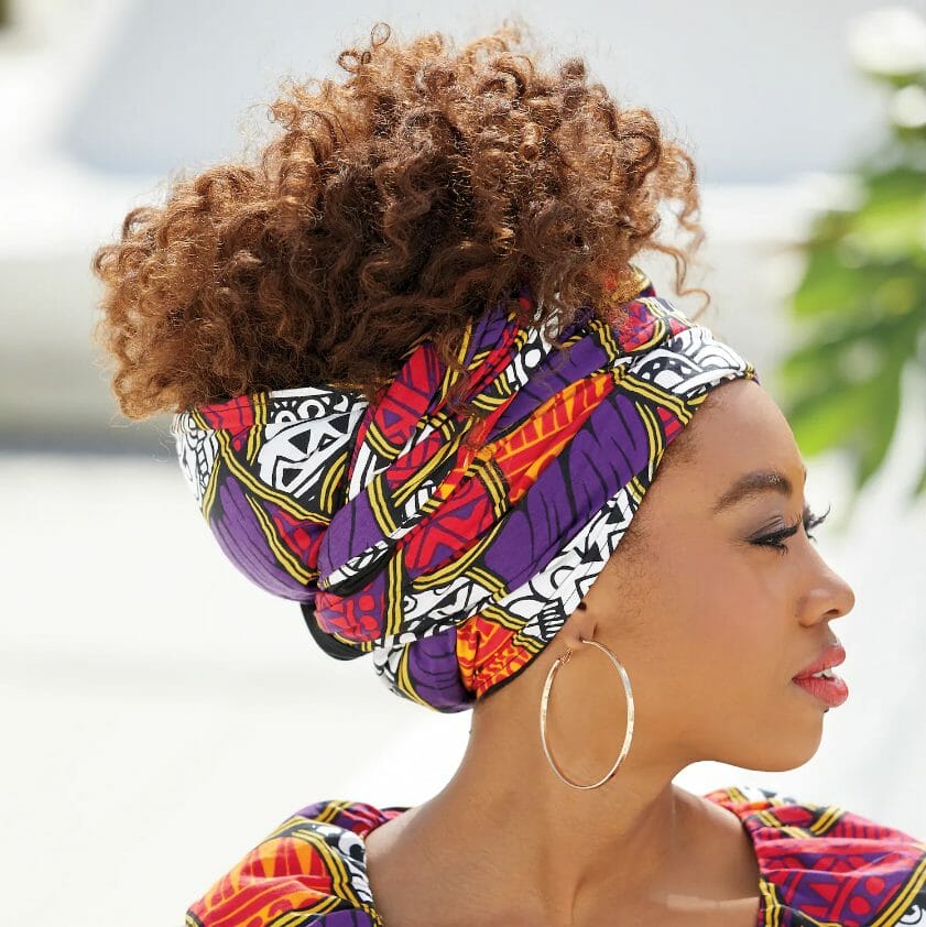 A profile view of an African-American woman wearing a purple, red and black print headwrap with hair piled high.