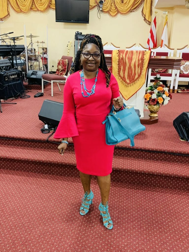 SHON, a smiling African-American woman in a hot pink dress with teal necklace, bag and shoes.