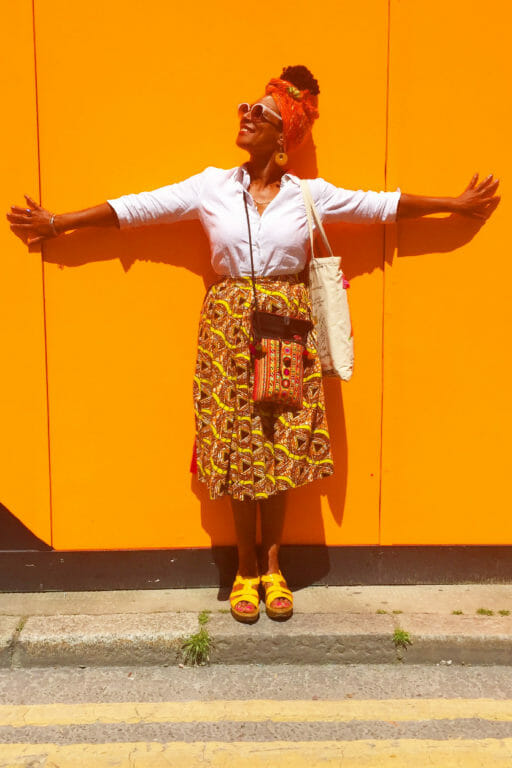 An African-American woman wearing a gold and red patterned dress, red headwrap, and yellow shoes standing with her arms stretched out against an orange wall.