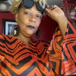 Woman posing in a orange and black pattern caftan with afrocentric earring and black sunglasses.